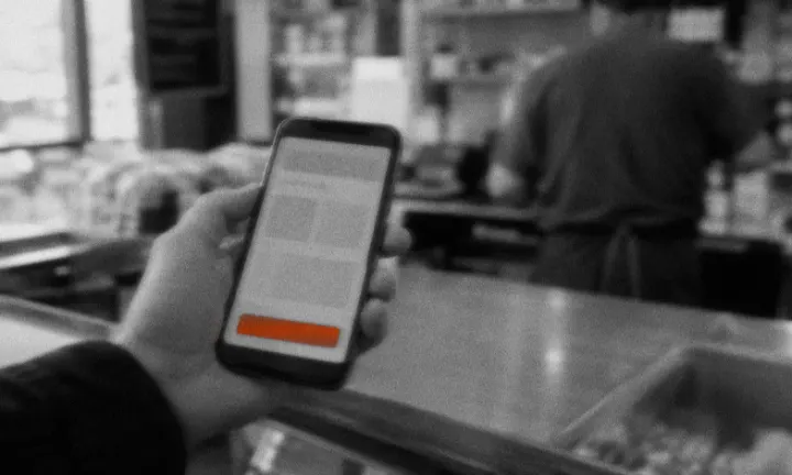 Mano con un celular que muestra una app con un botón, frente al mostrador de un negocio donde atienden