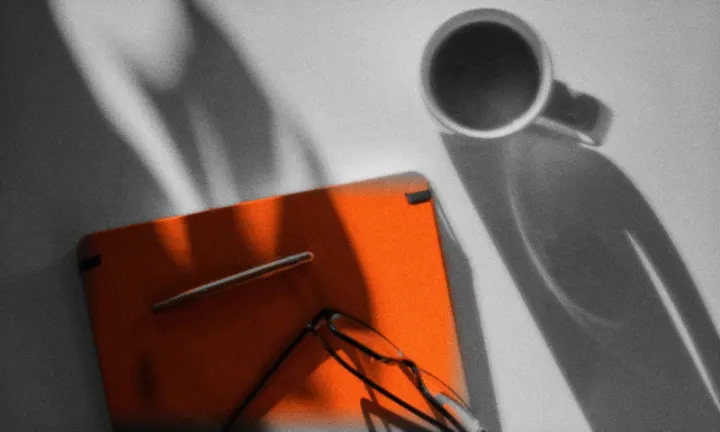 Cuaderno naranja con lapicera y anteojos sobre un escritorio, con sombras de ventana y una taza de café