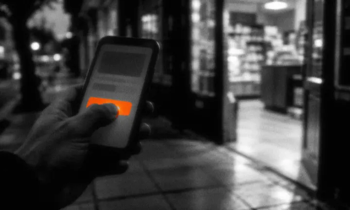 Mano con un celular de noche que toca un botón, frente a un local abierto y con luz